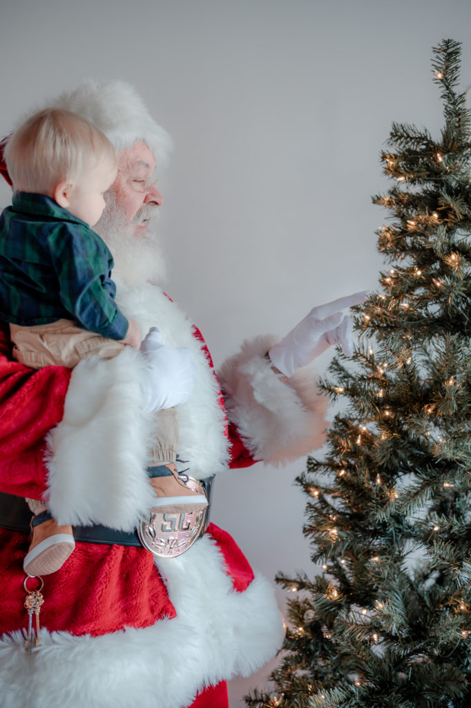 Santa Claus holding baby boy and pointing to  Christmas tree during 2020 Santa Mini photo session