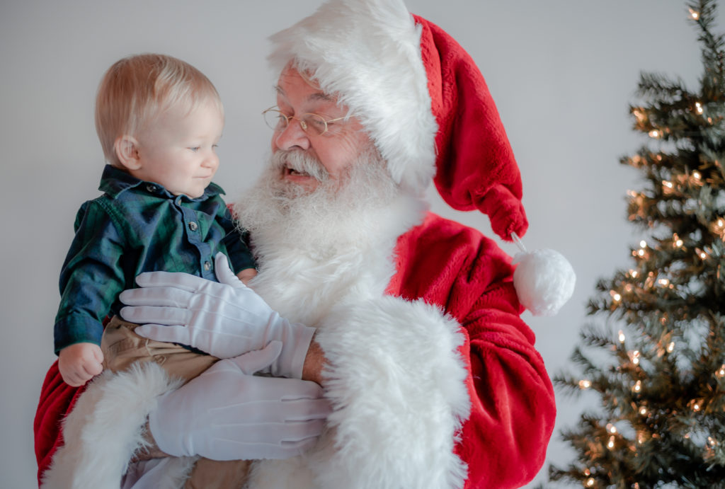 Santa Claus holding baby boy beside Christmas tree during 2020 Santa Mini photo session