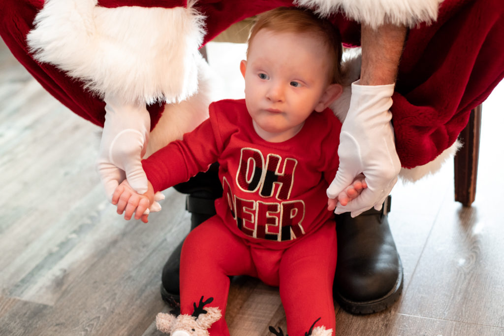 Santa holding baby's hands while baby is sitting on floor during a “Home for the Holidays” Santa Session