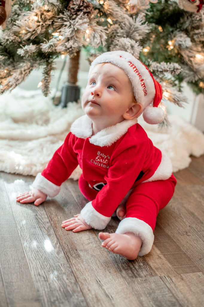 Baby boy in Christmas outfit sitting on floor looking up at tree during a “Home for the Holidays” Santa Session