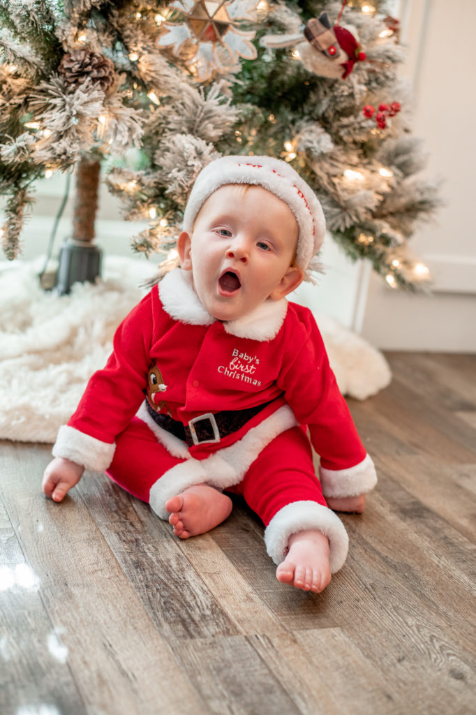 Baby boy in Santa suit, yawning, and sitting in front of Christmas tree during a “Home for the Holidays” Santa Session