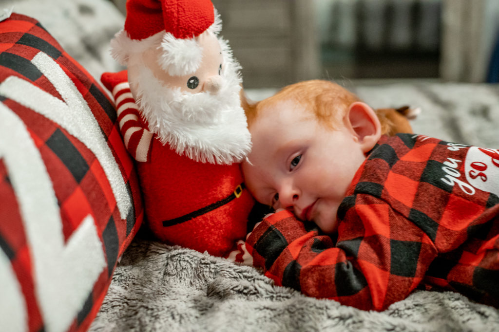 Baby boy in Christmas outfit falling asleep on bed during a “Home for the Holidays” Santa Session