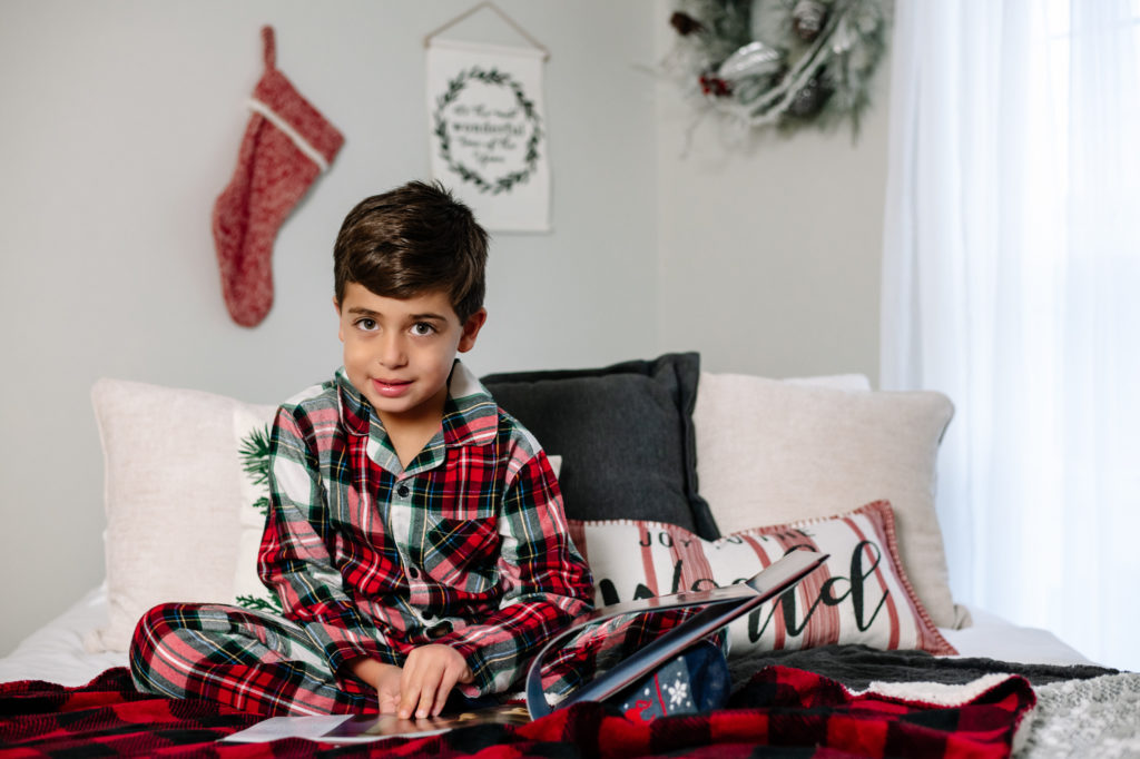 Young boy in Christmas pajamas on bed reading a book.